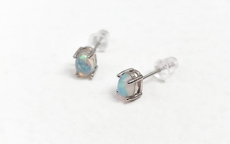 May include: A pair of stud earrings featuring oval-shaped opals set in silver-colored metal. The opals display a mix of pastel colors, including blue, pink, and white. The earrings have a post and backing for secure wear.