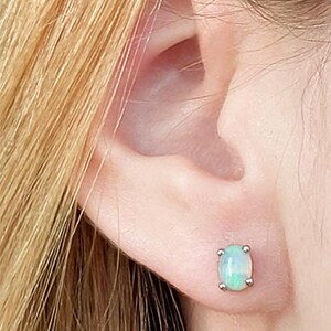 May include: A close-up of a person's ear with a small, square-shaped opal stud earring. The opal has a pale blue-green color and is set in a silver metal. The person's blonde hair is visible in the upper left corner.