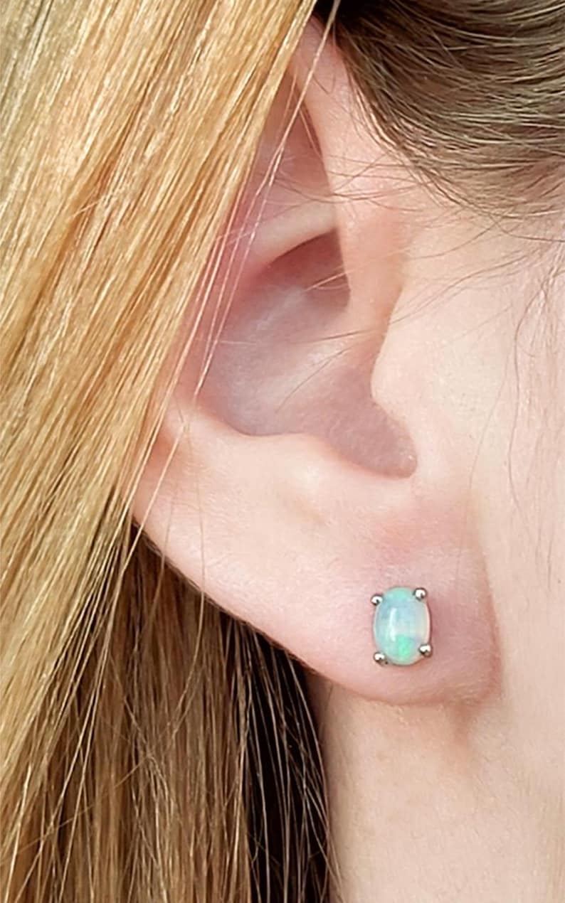 May include: A close-up of a person's ear with a small, square-shaped opal stud earring. The opal has a pale blue-green color and is set in a silver metal. The person's blonde hair is visible in the upper left corner.