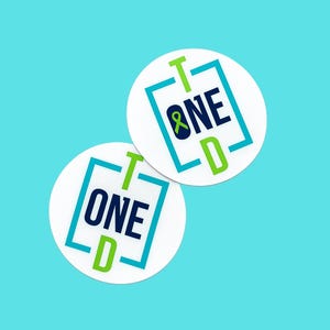 May include: Two circular white stickers featuring a graphic design with the text T1D ONE. The design includes a blue and green square border, with the letter T in green, the word ONE in dark blue, and the letter D in green. A small green ribbon is integrated into the letter O.