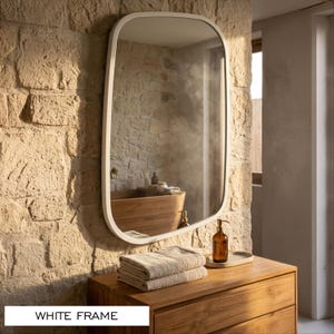 May include: A wall-mounted mirror featuring a smooth, rounded rectangular white frame. The reflective glass surface displays a clear view of a wooden vanity, a stack of folded beige towels, a brown glass soap dispenser, and a stone wall.
