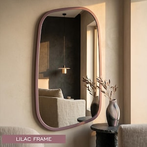 May include: A wall-mounted mirror with an irregular, rounded rectangular shape and a solid lilac frame. The reflective surface shows a room interior with a beige armchair, a hanging pendant light, and a vase with dried branches.