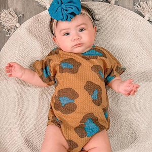Puede incluir: Un beb&eacute; con un body con estampado de leopardo marr&oacute;n y azul y una diadema con lazo turquesa. El beb&eacute; est&aacute; acostado sobre una alfombra redonda beige con borlas decorativas.