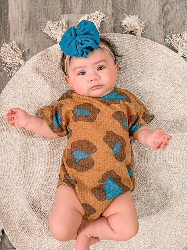 Puede incluir: Un beb&eacute; con un body con estampado de leopardo marr&oacute;n y azul y una diadema con lazo turquesa. El beb&eacute; est&aacute; acostado sobre una alfombra redonda beige con borlas decorativas.