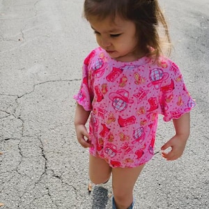 Puede incluir: Una ni&ntilde;a peque&ntilde;a con un body rosa y un lazo a juego con un estampado de tem&aacute;tica vaquera. La ni&ntilde;a tambi&eacute;n lleva botas azules con purpurina. El conjunto es perfecto para una fiesta tem&aacute;tica o para el d&iacute;a a d&iacute;a.
