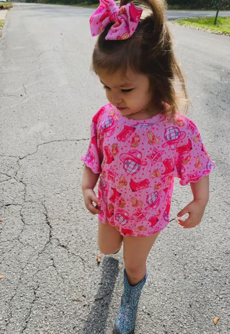 Puede incluir: Una ni&ntilde;a peque&ntilde;a con un body rosa y un lazo a juego con un estampado de tem&aacute;tica vaquera. La ni&ntilde;a tambi&eacute;n lleva botas azules con purpurina. El conjunto es perfecto para una fiesta tem&aacute;tica o para el d&iacute;a a d&iacute;a.
