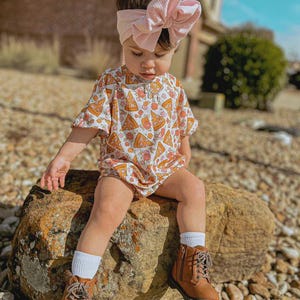 Puede incluir: Un beb&eacute; con una diadema de lazo rosa claro, un pelele de manga corta con un estampado de porciones de pizza, calcetines blancos y botas marrones con cordones. El ni&ntilde;o est&aacute; sentado en una gran roca al aire libre.