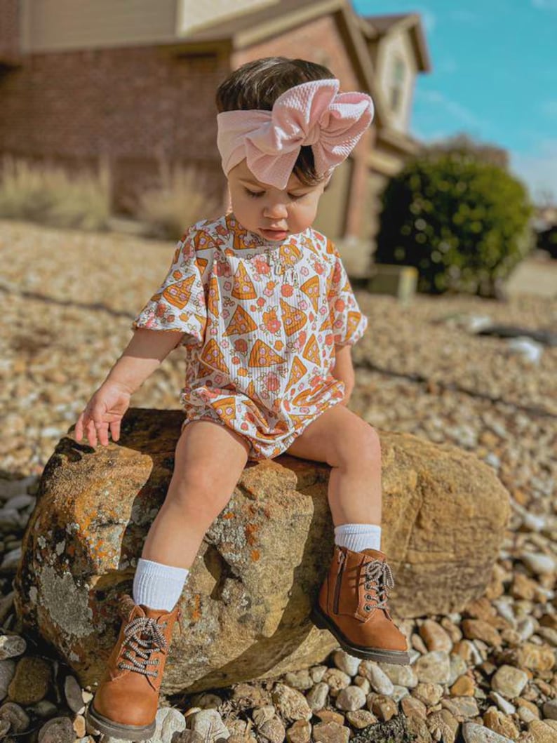 Puede incluir: Un beb&eacute; con una diadema de lazo rosa claro, un pelele de manga corta con un estampado de porciones de pizza, calcetines blancos y botas marrones con cordones. El ni&ntilde;o est&aacute; sentado en una gran roca al aire libre.