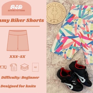 May include: A sewing pattern for biker shorts titled Mommy Biker Shorts with a size range of XXS-5X. The image displays a pair of white biker shorts featuring a colorful abstract pattern of pink, blue, and yellow brushstrokes, alongside black cycling shoes.