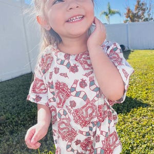 Puede incluir: Un pelele blanco con un estampado rojo y verde con la palabra "Slytherin" y un dise&ntilde;o de serpiente. El pelele tiene mangas cortas y cuello redondo. El ni&ntilde;o sonr&iacute;e y mira hacia arriba.