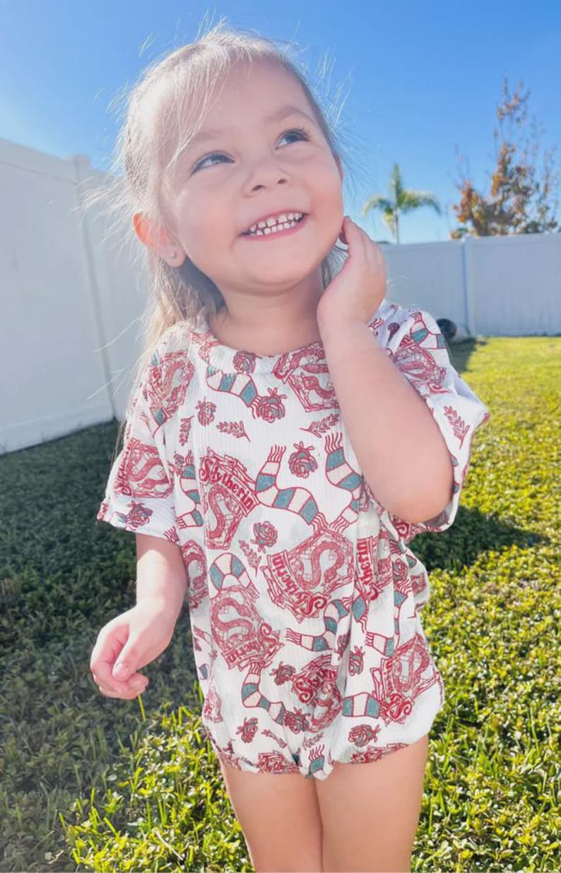 Puede incluir: Un pelele blanco con un estampado rojo y verde con la palabra "Slytherin" y un dise&ntilde;o de serpiente. El pelele tiene mangas cortas y cuello redondo. El ni&ntilde;o sonr&iacute;e y mira hacia arriba.