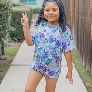Puede incluir: Una ni&ntilde;a peque&ntilde;a con un lazo azul, un body de manga corta con un dise&ntilde;o de dibujos animados y pantalones cortos. El body presenta un estampado colorido con varios personajes de dibujos animados. La ni&ntilde;a sonr&iacute;e y hace el signo de la paz.