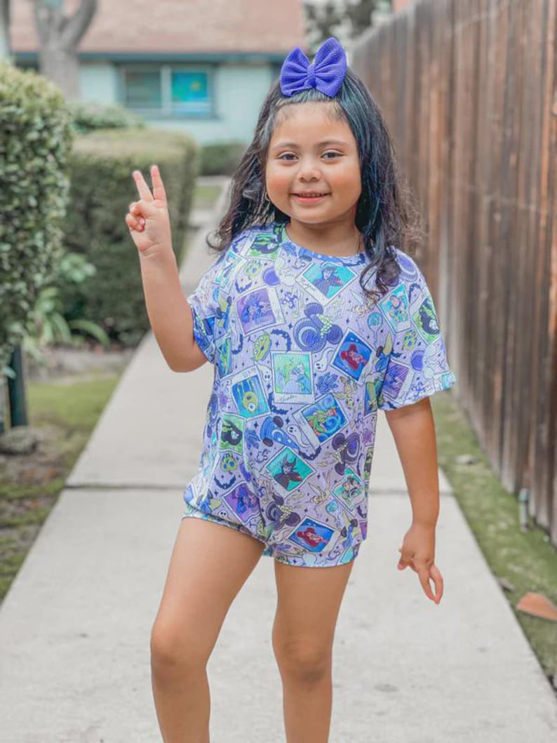 Puede incluir: Una ni&ntilde;a peque&ntilde;a con un lazo azul, un body de manga corta con un dise&ntilde;o de dibujos animados y pantalones cortos. El body presenta un estampado colorido con varios personajes de dibujos animados. La ni&ntilde;a sonr&iacute;e y hace el signo de la paz.