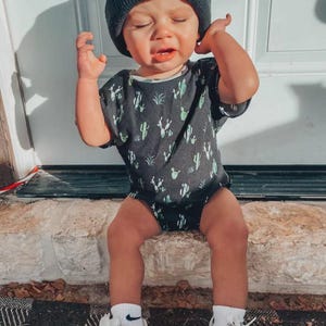 Puede incluir: Un ni&ntilde;o peque&ntilde;o con un gorro negro de Adidas, un body gris oscuro con estampado de cactus, calcetines blancos y zapatillas Adidas blancas con rayas negras. El ni&ntilde;o est&aacute; sentado en un borde de piedra.