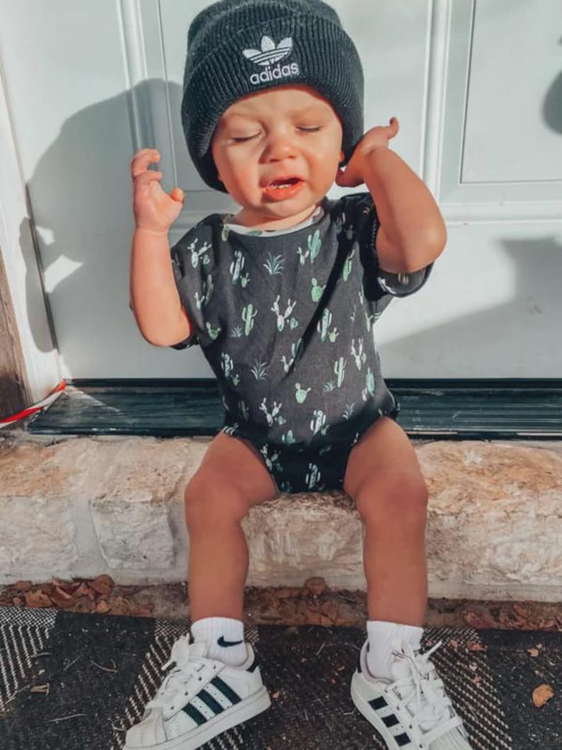 Puede incluir: Un ni&ntilde;o peque&ntilde;o con un gorro negro de Adidas, un body gris oscuro con estampado de cactus, calcetines blancos y zapatillas Adidas blancas con rayas negras. El ni&ntilde;o est&aacute; sentado en un borde de piedra.
