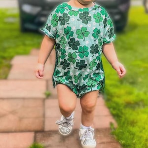 Puede incluir: Un ni&ntilde;o peque&ntilde;o con un pelele verde con estampado de tr&eacute;boles y flecos. El ni&ntilde;o tambi&eacute;n lleva gafas de sol, un lazo para el pelo y zapatillas brillantes. La imagen est&aacute; tomada al aire libre en un camino de ladrillos.