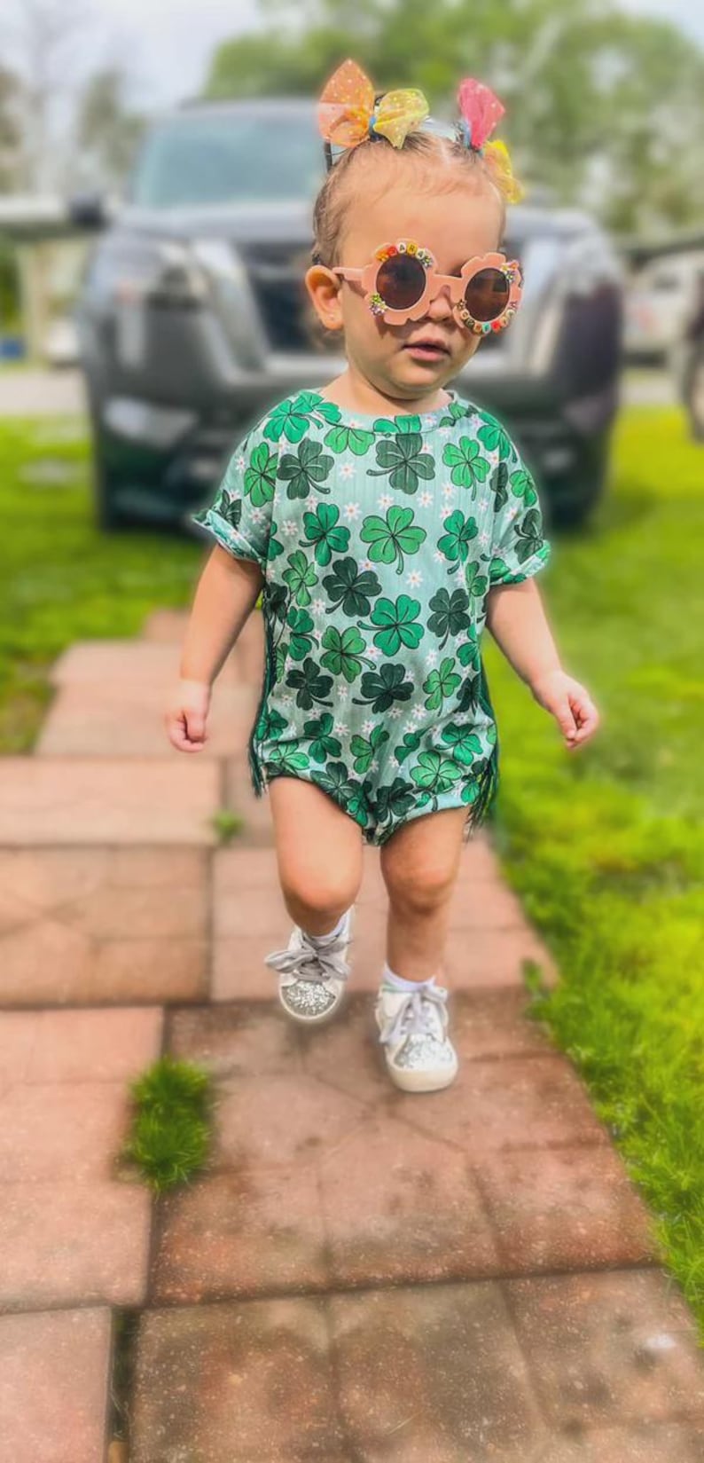 Puede incluir: Un ni&ntilde;o peque&ntilde;o con un pelele verde con estampado de tr&eacute;boles y flecos. El ni&ntilde;o tambi&eacute;n lleva gafas de sol, un lazo para el pelo y zapatillas brillantes. La imagen est&aacute; tomada al aire libre en un camino de ladrillos.