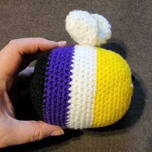 May include: A crocheted bee plush toy with black, purple, white and yellow stripes. The bee has white crocheted wings.