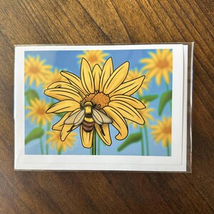 May include: A set of greeting cards featuring a vibrant illustration of a bee on a yellow sunflower against a blue sky background. The cards are in a clear plastic sleeve, perfect for gifting or personal use.