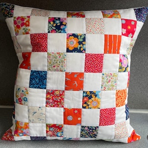 May include: A square patchwork cushion with a white base and a grid of colourful floral and patterned squares. The squares feature designs in red, orange, blue, and green. The cushion has a white border.