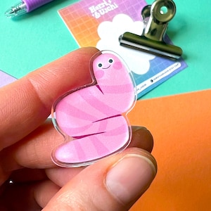 Könnte beinhalten: Ein rosa-weiß gestreifter Wurm mit einem lächelnden Gesicht aus klarem Acryl wird in einer Hand gehalten. Der Wurm ist ein süßes und skurriles Design.