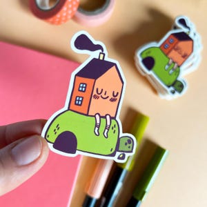 Pode incluir: Um adesivo de uma casa de desenho animado com uma chaminé e um rosto sorridente sentado em uma tartaruga verde com uma concha. A casa é laranja e tem duas janelas. A tartaruga é verde e tem uma concha marrom.