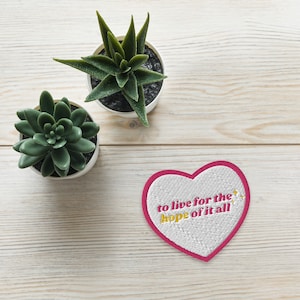 Könnte beinhalten: Ein herzförmiger Aufnäher mit rosa Rand und dem gestickten Text "to live for the hope of it all" in Gelb und Rosa. Zwei kleine Sukkulenten in Töpfen befinden sich ebenfalls im Bild, vor einem hellen Holzmaserungshintergrund.
