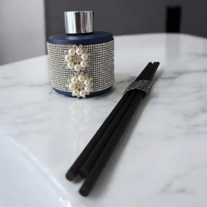 May include: A dark blue glass diffuser bottle with a silver lid and a decorative design of white pearls. The bottle is sitting on a white marble surface with black diffuser sticks.