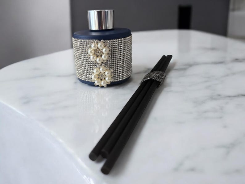 May include: A dark blue glass diffuser bottle with a silver lid and a decorative design of white pearls. The bottle is sitting on a white marble surface with black diffuser sticks.