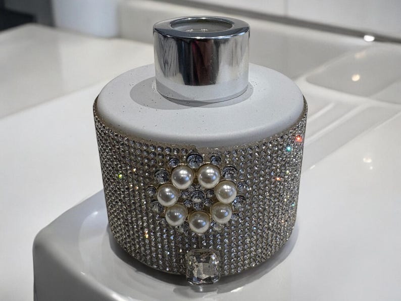 May include: A white ceramic diffuser bottle with a silver lid, covered in sparkling rhinestones and a pearl-like embellishment.