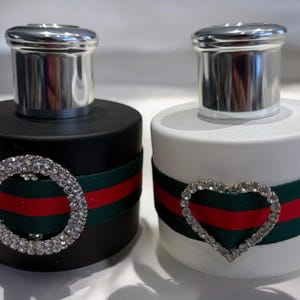 Könnte beinhalten: Zwei Parfümflakons mit silbernen Kappen und einem roten, grünen und weißen Streifenband um die Mitte. Eine Flasche ist schwarz mit einem Strassstein-Kreisdesign und die andere ist weiß mit einem Strassstein-Herzdesign.