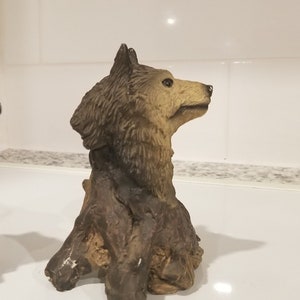 May include: A brown and tan wolf figurine sitting on a tree stump. The wolf is looking to the right and has a detailed fur texture.