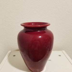 May include: A glossy, deep red ceramic vase with a rounded body and a slightly flared rim. The vase is displayed on a white surface against a plain wall. The vase is approximately 25 cm tall.