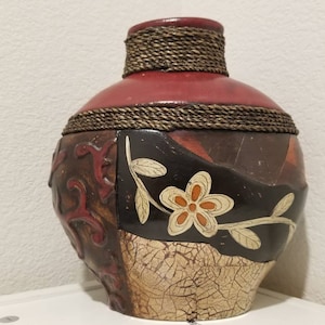 Puede incluir: Un jarrón de cerámica redondo de color rojo, marrón y crema con un diseño de cuerda alrededor de la parte superior e inferior. El jarrón tiene un diseño floral con flores blancas, naranjas y amarillas.