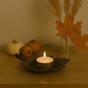 Op de afbeelding: Een brandende waxinelichtkaars in een bladvormige houder. De kaars is omgeven door kleine pompoenen en herfstbladeren, waaronder oranje esdoornbladeren in een glazen vaas. De scène creëert een gezellige herfstsfeer.