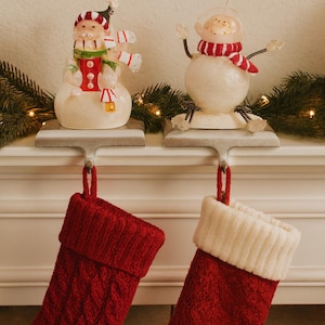 May include: Two Christmas stocking holders with decorative snowmen and red stockings. One snowman wears a hat and scarf, the other has a space helmet and scarf. The red stockings have a cable knit design.