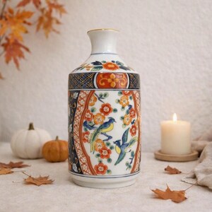May include: A ceramic vase featuring a white base with intricate patterns of orange flowers and blue birds. The design includes dark blue panels with gold accents and a narrow neck. The surface is decorated with detailed botanical and avian motifs.