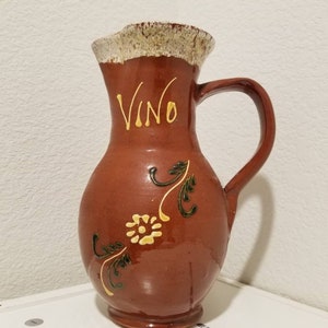 May include: A brown ceramic jug with a white rim and a handle. The jug has a floral design and the word "Vino" written on the front.