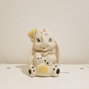 May include: A ceramic bunny-shaped money box in off-white with gold detailing. The bunny has a yellow hat with a dollar sign and black eyes and paws. The money box has a slot on top for inserting coins.