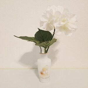 May include: White ceramic vase with a gold rim and a floral design featuring a couple of angels. The vase is filled with a white hydrangea with green leaves.