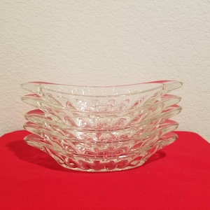 Puede incluir: Una pila de cinco cuencos de vidrio transparente para banana split. Cada cuenco tiene una forma curva, similar a un barco, con un diseño texturizado y estampado. Los cuencos están apilados sobre una superficie roja contra una pared blanca.