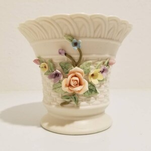 Puede incluir: Un jarrón de cerámica blanco con un diseño de tejido de cesta y detalles florales en 3D. Las flores son rosas, amarillas, azules y moradas.