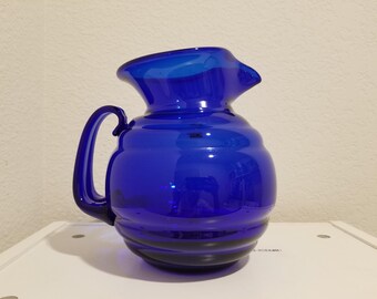 May include: A cobalt blue glass pitcher with a rounded body, a curved handle, and a spout. The pitcher has a glossy finish and sits on a white surface. The design features horizontal ridges around the body.