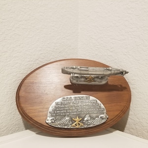 May include: A wooden plaque with a metal submarine model and an engraved metal plate. The plate reads "C.S.S. HUNLEY THE FIRST TRUE SUBMARINE" with additional text. The submarine model is silver-colored.