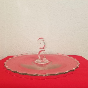 Puede incluir: Un soporte para pasteles de vidrio transparente con un borde festoneado decorativo y un asa curva. El soporte está sobre una superficie roja.