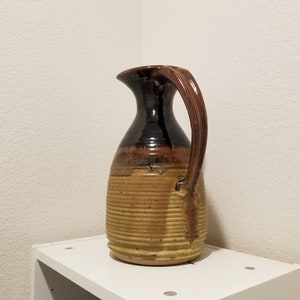 Puede incluir: Una jarra de cerámica marrón y amarilla con una boca ancha y un asa. La jarra tiene una superficie texturizada y un acabado brillante.