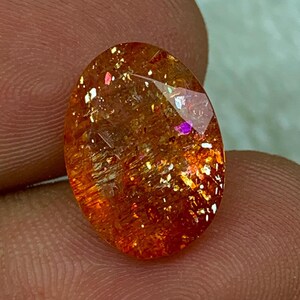 May include: A large, oval-shaped, orange gemstone with a glittery, iridescent finish. The stone has a reddish-orange hue with flecks of gold and silver.