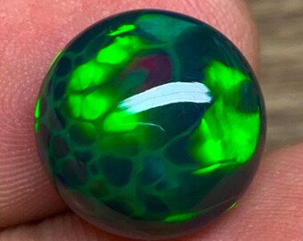 May include: A round, polished black opal gemstone with vibrant green and blue internal fire. The gemstone is held in a person's hand, showcasing its reflective qualities and intricate patterns. The opal's surface is smooth and glossy.