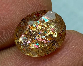 May include: A golden oval-shaped gemstone with iridescent inclusions. The gemstone has a faceted cut and is set in a clear, transparent setting.