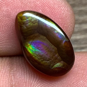 May include: A polished, oval-shaped gemstone with a dark brown base color. The stone displays iridescent colors, including green, purple, and blue, creating a shimmering effect. The gemstone is held between fingers.